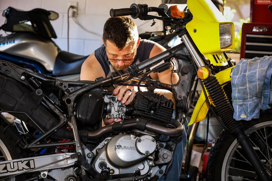 A mechanic intensely focused on repairing a motorcycle engine in a cluttered garage setting.